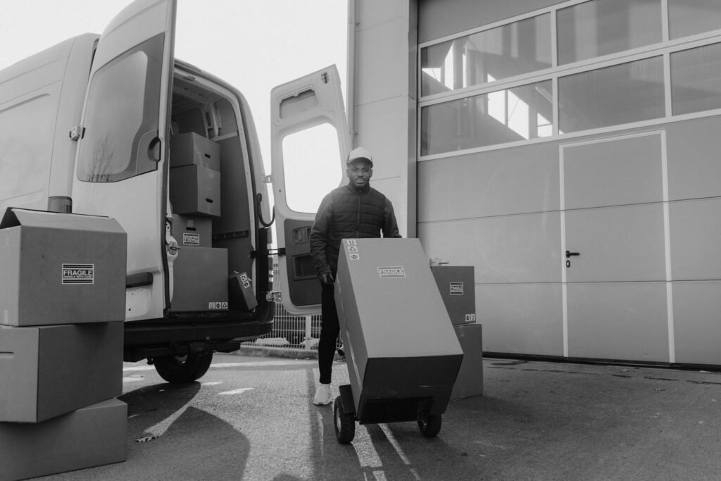 Monochrome image of a delivery man with boxes on a trolley near a van.